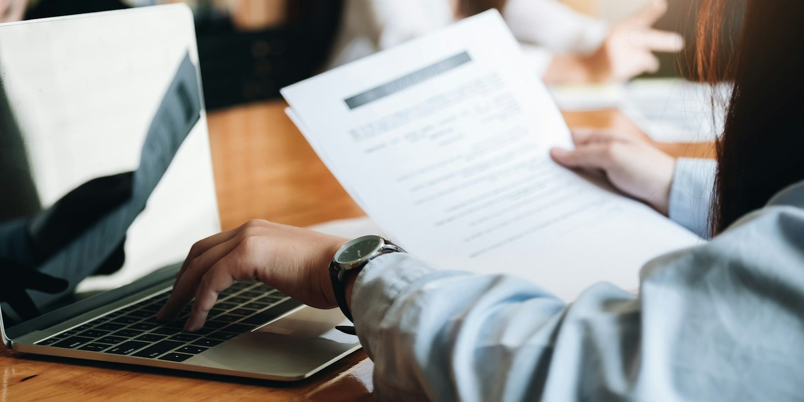 Eine Hand hält und liest ein Dokument neben einem Laptop auf einem Tisch.